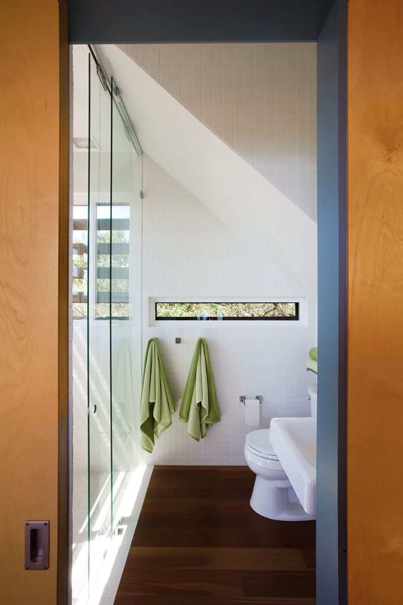 In this modern white and wood bathroom, small windows provide views and add natural light, however they are small enough to provide privacy at the same time. Floor-to-ceiling white tiles help to keep the interior bright. #ModernBathroom #WhiteBathroom #WoodFlooring #BathroomDesign