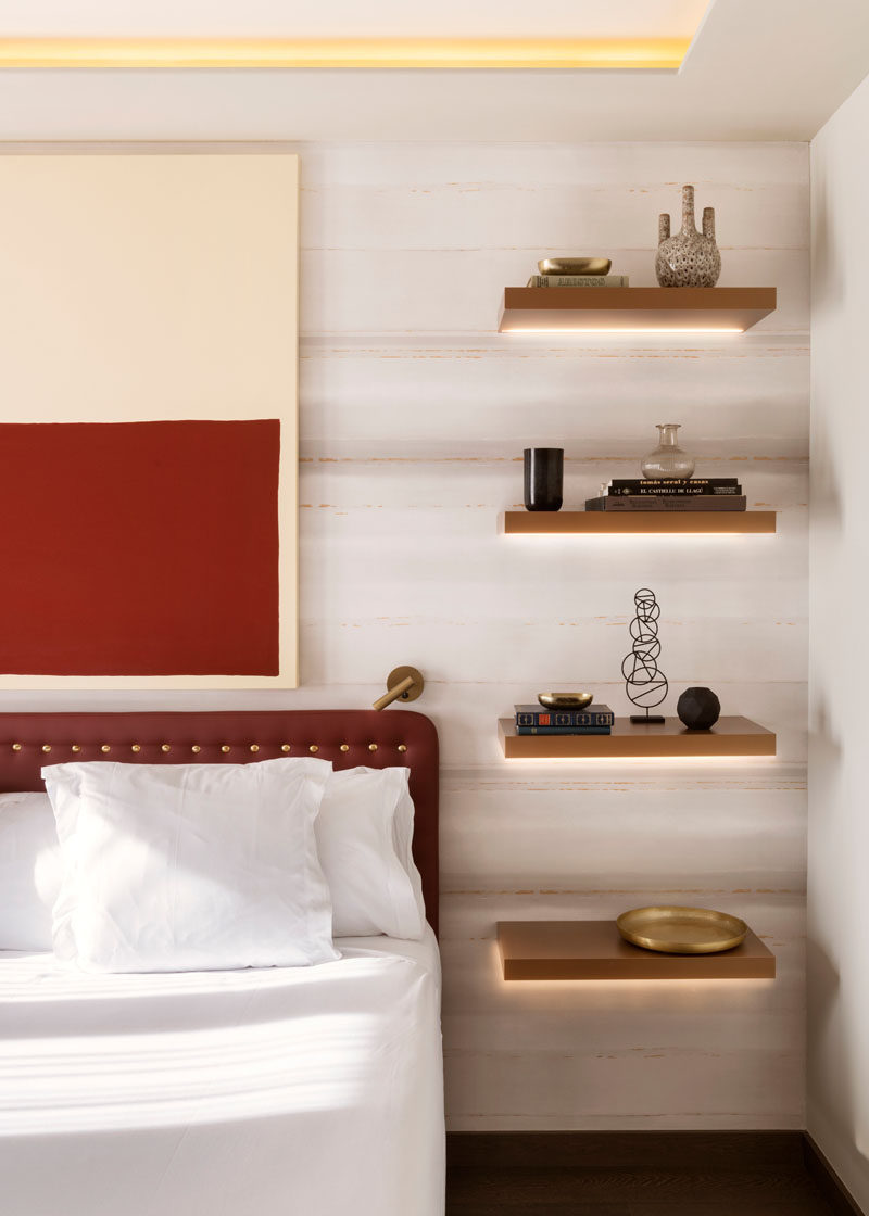 In this modern bedroom, four floating wood shelves with hidden lighting have been installed, with the lowest shelf at the correct height for a bedside table, while the other three shelves have been used to display decorative items. #BedroomDesign #BedsideTable #InteriorDesign #Shelving