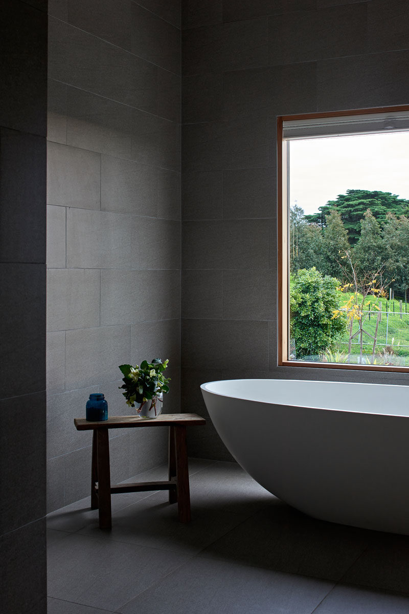 In this modern bathroom, large grey tiles have been paired with wood window frames and a white bathtub for a contemporary appearance. #GreyBathroom #GreyTiles #ModernBathroom #BathroomDesign