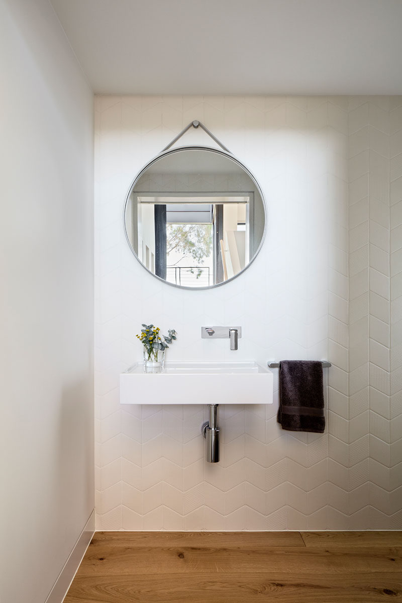 In this modern bathroom, a round mirror hangs on the wall, while white tiles in a chevron pattern cover the wall. #BathroomDesign #RoundMirror #ChevronTiles #WhiteTiles