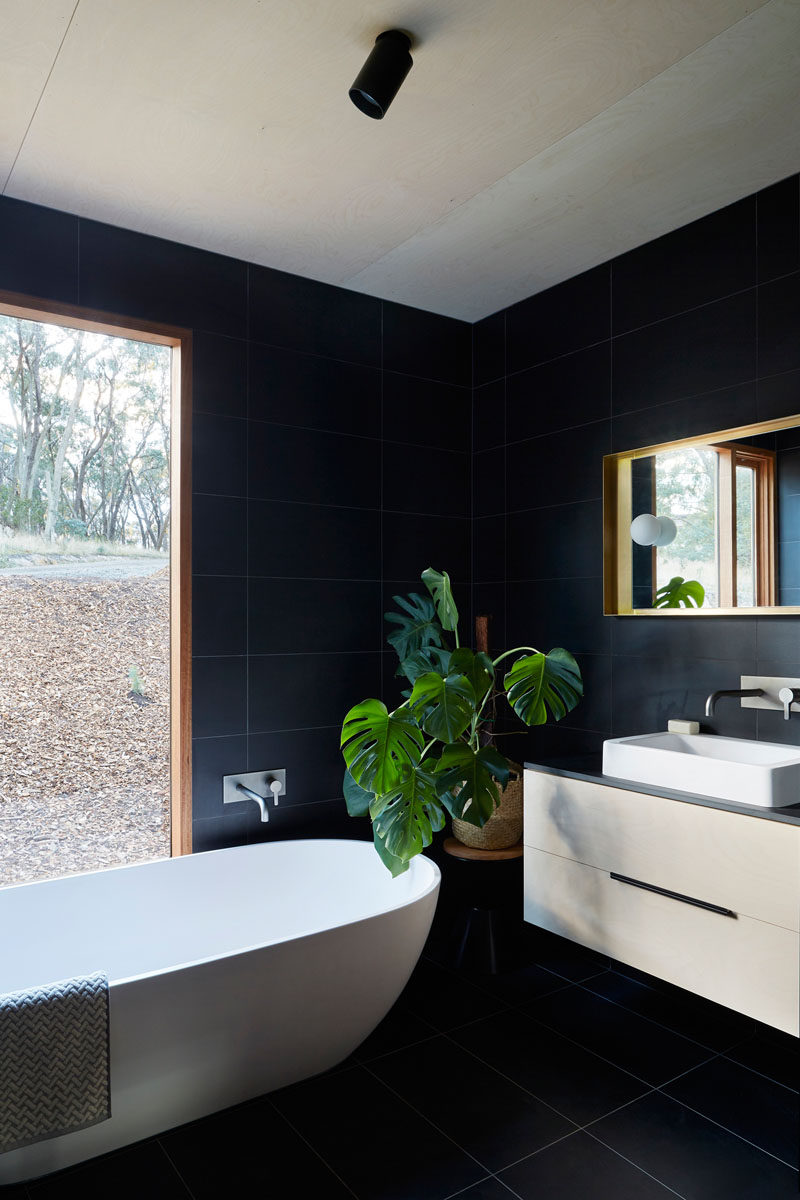 In this modern bathroom, large black tiles have been used on the walls and floor and they contrast the wood frames and white accents like the freestanding bathtub and sink. #BlackBathroom #ModernBathroom #BathroomDesign