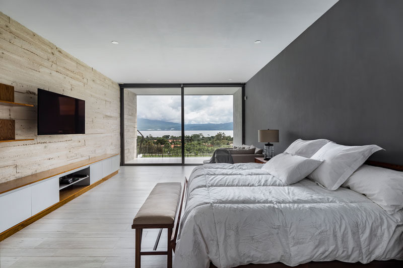 In this modern bedroom a board formed concrete wall sits opposite a dark grey accent wall, and both of them flow through to the exterior of the house. A sliding glass door opens the bedroom up to a private balcony with views of the neighborhood. #ConcreteWall #ModernBedroom #BedroomDesign