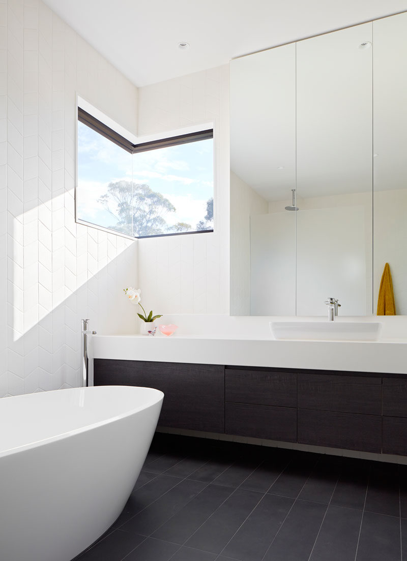 In this modern en-suite bathroom, a corner window adds natural light and provides a view of the trees, while dark flooring and a dark vanity contrast the white bathtub and white floor-to-ceiling tiles. #ModernBathroom #BathroomDesign #CornerWindow