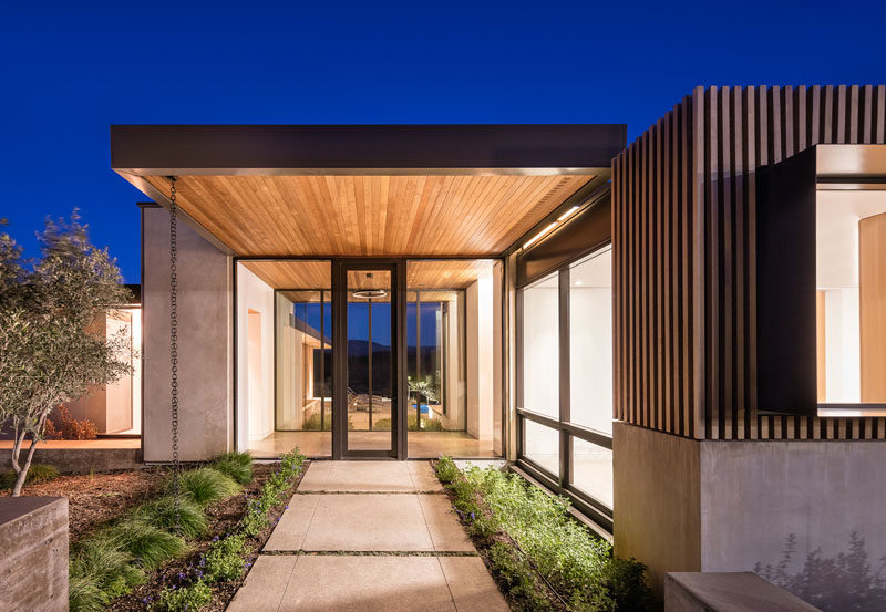 This modern house features large overhanging, wood lined roof sections that cover walkways and connect the various areas of the house. #ModernHouse #Roof #WoodOverhang #Front Door #Landscaping