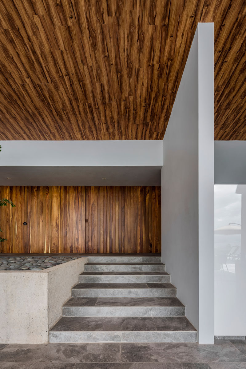 Stepping inside this modern house, there's an immediate warmth from the use of wood throughout the interior. A set of small stairs leads from the wood front door down into the open living area. #ModernHouse #WoodCeiling #WoodFrontDoor #InteriorSteps