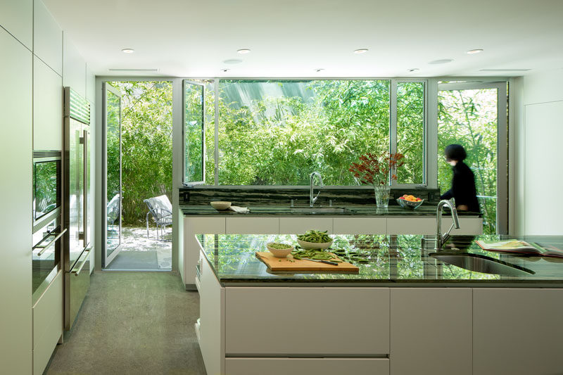 This minimalist white kitchen with dark countertops has views of the garden, and two floor-to-ceiling glass doors can be opened to a small patio area. #MinimalistKitchen #WhiteCabinets #ModernKitchen