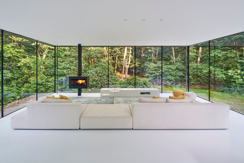 Inside this modern house, the floor and ceiling, as well as the furnishings have been kept bright, allowing natural light to reflect throughout the interior. #WhiteInterior #GlassWalls #ModernLivingRoom #Fireplace #Windows