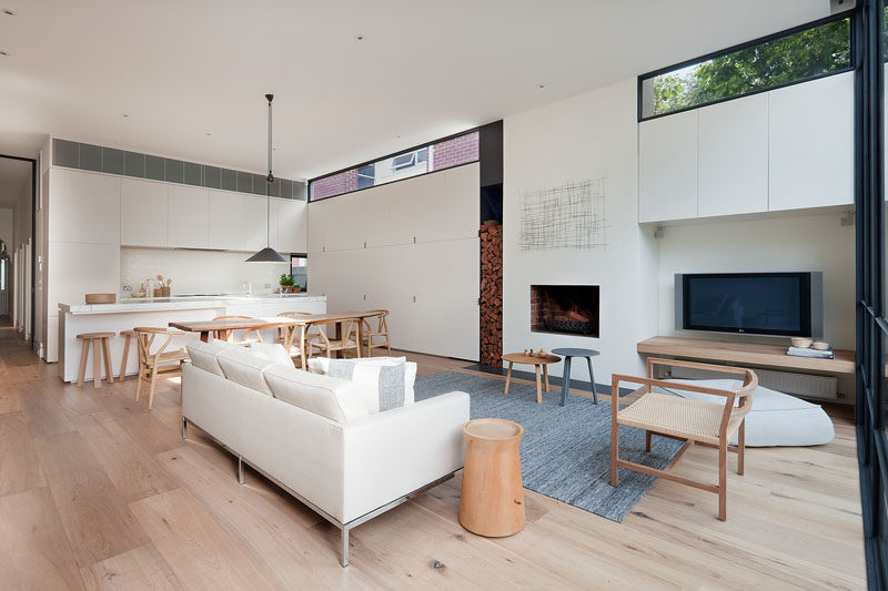 This modern open plan interior has a fireplace and wood storage that sits flush with the minimalist white cabinets in the kitchen. #WhiteKitchenCabinets #ModernLivingRoom #ModernFireplace #WoodStorage 