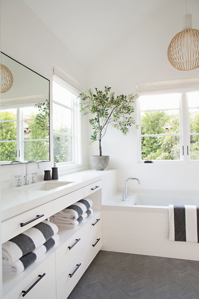 In this white and contemporary bathroom, a custom white vanity with open storage and built-in drawers, runs the length of the wall and drops down to create a built-in bathtub, while grey floor tiles in a herringbone pattern and multiple windows break up the all white room. #WhiteBathroom #ContemporaryBathroom #BuiltInBath #BathroomDesign