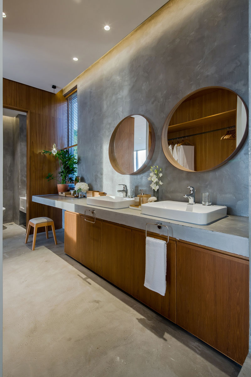 This modern bathroom has a large wood vanity with a concrete countertop that matches the floor. A round wood-framed mirror hangs above each sink, while the vanity extends to create additional counter space with an open space for a stool. #ModernBathroom #WoodAndConcreteBathroom #RoundMirrors #ConcreteCountertop