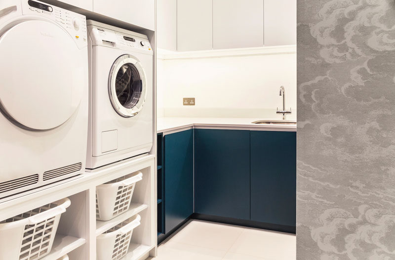 This modern utility room features white and dark blue matte cabinetry, a washer and dryer that have been raised so there's no need to bend down when moving the washing to the dryer. #ModernLaundry #LaundryRoom