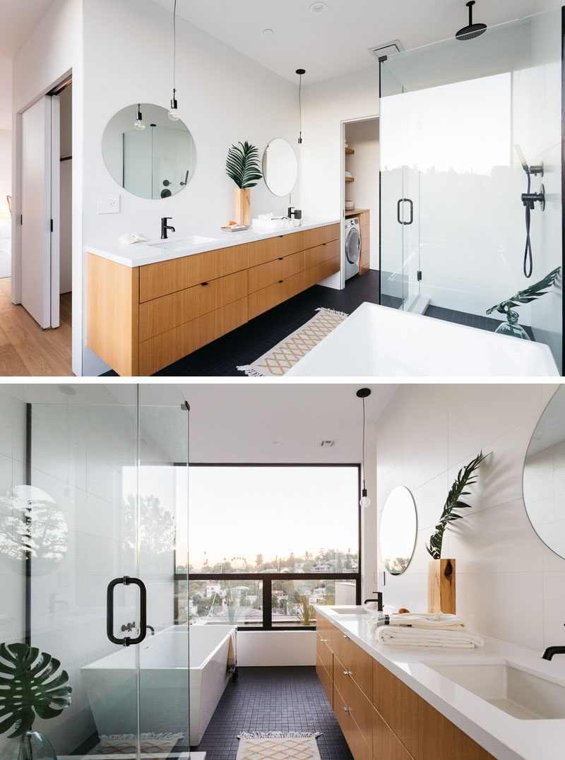 In this modern master bathroom, there's a large wood vanity with a white counter and round mirrors. Beside the vanity is a small nook with a laundry room, and opposite the vanity is the shower with a glass surround and a bathtub positioned to take advantage of the views. #MasterBathroom #BathroomDesign #ModernBathroom