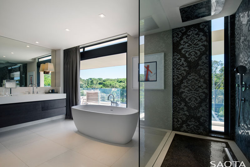 Decorative black tiles have been used in the shower of this bathroom, while a white standalone bathtub is positioned to take in tree views. The vanity combines both black cabinets and a white countertop to complement the overall contemporary look. #ModernBathroom #BlackAndWhite