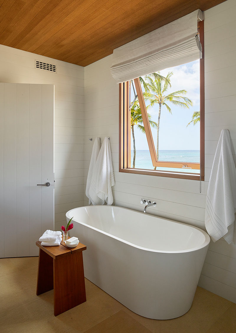 In this modern beach house bathroom, a freestanding white bathtub sits underneath a window with views of the beach. #ModernBathroom #WhiteAndWood