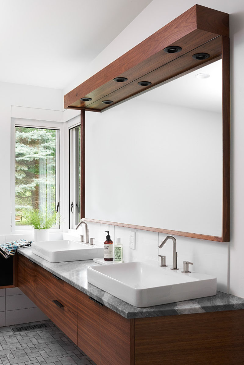 In this modern master bathroom, a dark wood-framed mirror sits above the vanity with dual sinks and a stone counter. #MasterBathroom #BathroomDesign