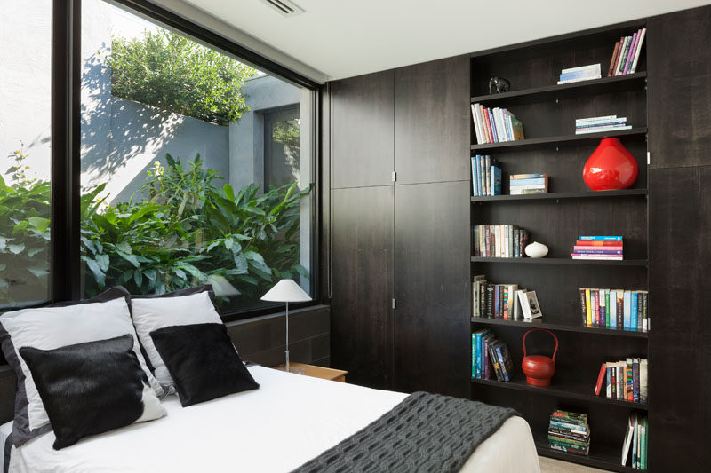 In this modern bedroom, dark wood shelving and closets line the wall, while large windows provide a glimpse of the lush plants outside. #ModernBedroom #Shelving #Windows