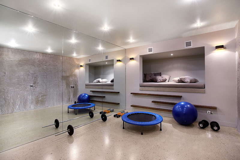 This modern house has a multi-purpose room that's been set up as a gym. A mirrored wall for when working out, or when relaxing, someone can cozy up in the raised day bed that's built into the wall. #HomeGym #BuiltInDayBed