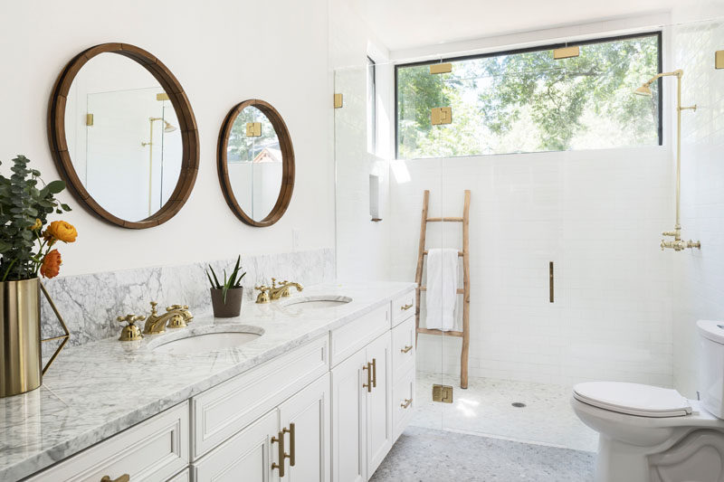 In this contemporary bathroom, round mirrors hang above the double-sink vanity that's topped with Carrara marble, while a glass shower surround allows the natural light from the window to flow through the space. #BathroomDesign #WhiteBathroom