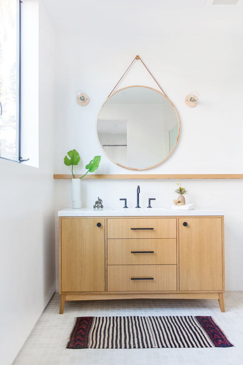 This modern bathroom features a round mirror hanging on the wall, a simple wood shelf and a wood vanity with matte black hardware. #BathroomDesign #ModernBathroom