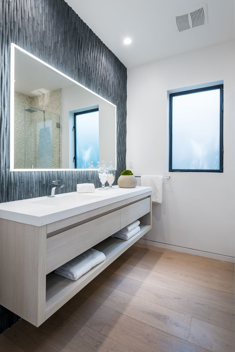 In this modern bathroom three dimensional grey tiles have been used to create an accent wall, while a backlit mirror highlights the design of the wall. #ModernBathroom #BathroomAccentWall #BacklitMirror