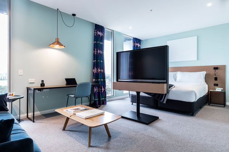 This modern hotel room has a television stand separating the bedroom space from the living space, while a desk is positioned against the wall under a single copper pendant light. #HotelRoom #InteriorDesign