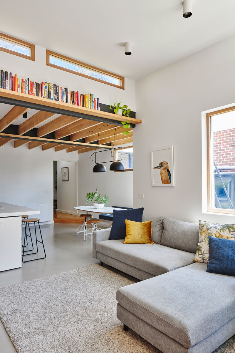 This modern living room has been set up with a simple grey couch and matching rug. A wood book shelf that's affixed to a beam draws the eye upwards to the height of the room and the windows. #LivingRoom #Shelving