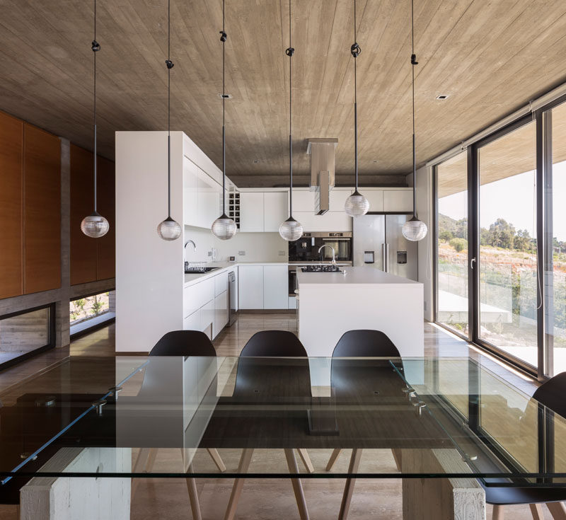 This glass, steel and concrete dining table is anchored in the room by the use of 6 pendant lights that hang from the ceiling. Click through to see more photos of this concrete house. #DiningRoom #InteriorDesign #WhiteKitchen