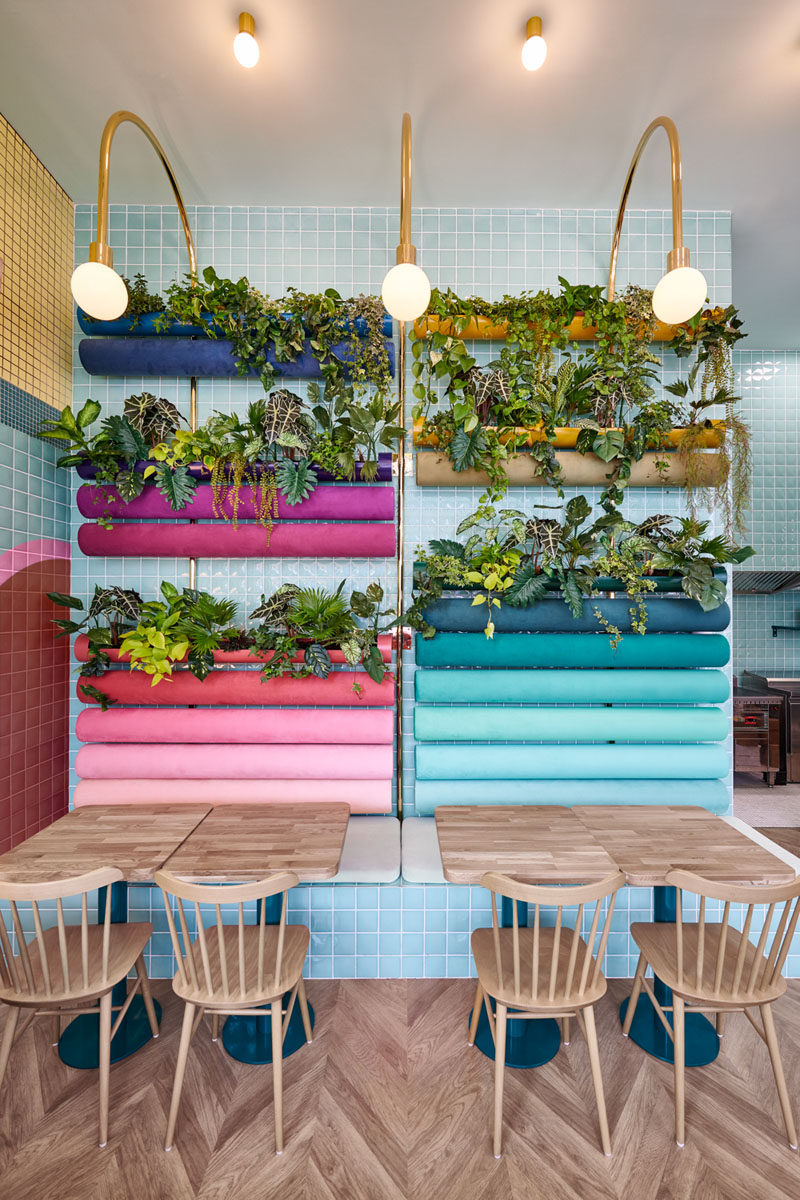 In this modern restaurant, plants have been added to the back 'rainbow' cushions on the wall in the seating area, while gold bar lamps curve over to add light to the space and tie in with the customer service area. Click through to see more photos of this interior. #RestaurantDesign #Planters #Seating #InteriorDesign