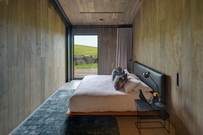 In this modern guest bedroom, there's wood walls and ceilings, a concrete floor with a blue rug, and a glass door that frames the view and opens to a small wood deck. Click through to see more photos of this modern guest house. #GuestHouse #Bedroom #WoodWalls #GlassDoor #ConcreteFloor #GuestBedroom