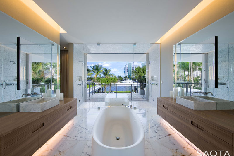 In this modern master bathroom, wood vanities have been placed on either side of the room with a freestanding bathtub between them, while the glass enclosed showers have water views. #ModernBathroom