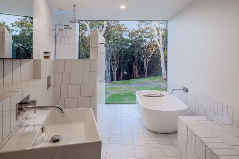 This modern white bathroom has a freestanding bathtub, rectangular white tiles, and a large picture window that frames the view. Click through to see more photos of this modern house. #WhiteBathroom #ModernBathroom #Bathroom #WhiteTiles