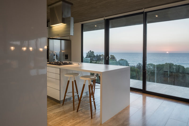 This bright white kitchen has an island and views of the water through floor-to-ceiling windows and a glass door. Click through to see more photos of this modern house. #Kitchen #WhiteIsland #Windows