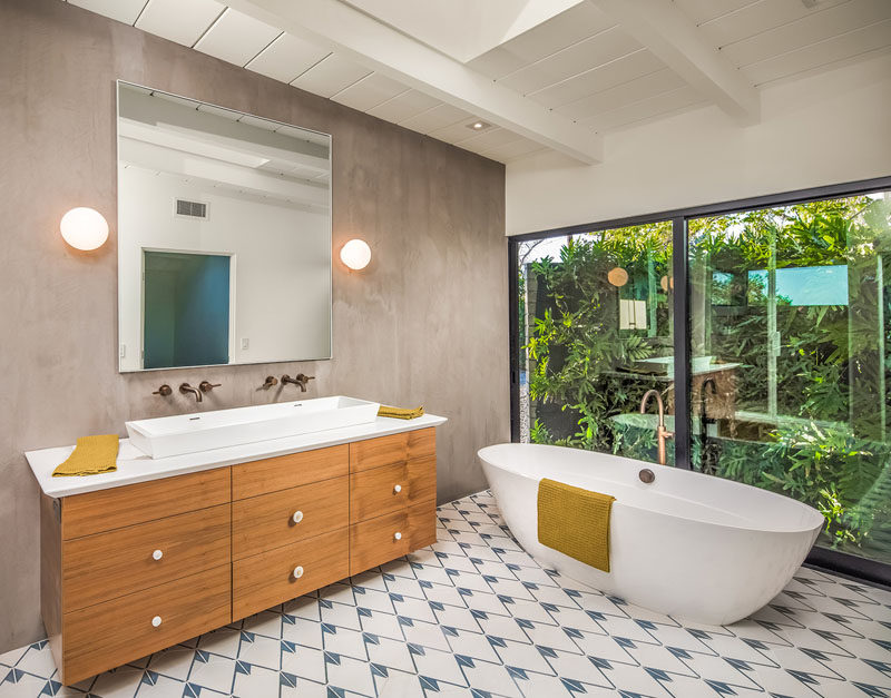 In this ensuite bathroom, there's a standalone bathtub that takes advantage of the large floor-to-ceiling windows, and has a view of a lush garden. #Bathroom #Ensuite #WoodVanity #Windows #StandaloneBathtub