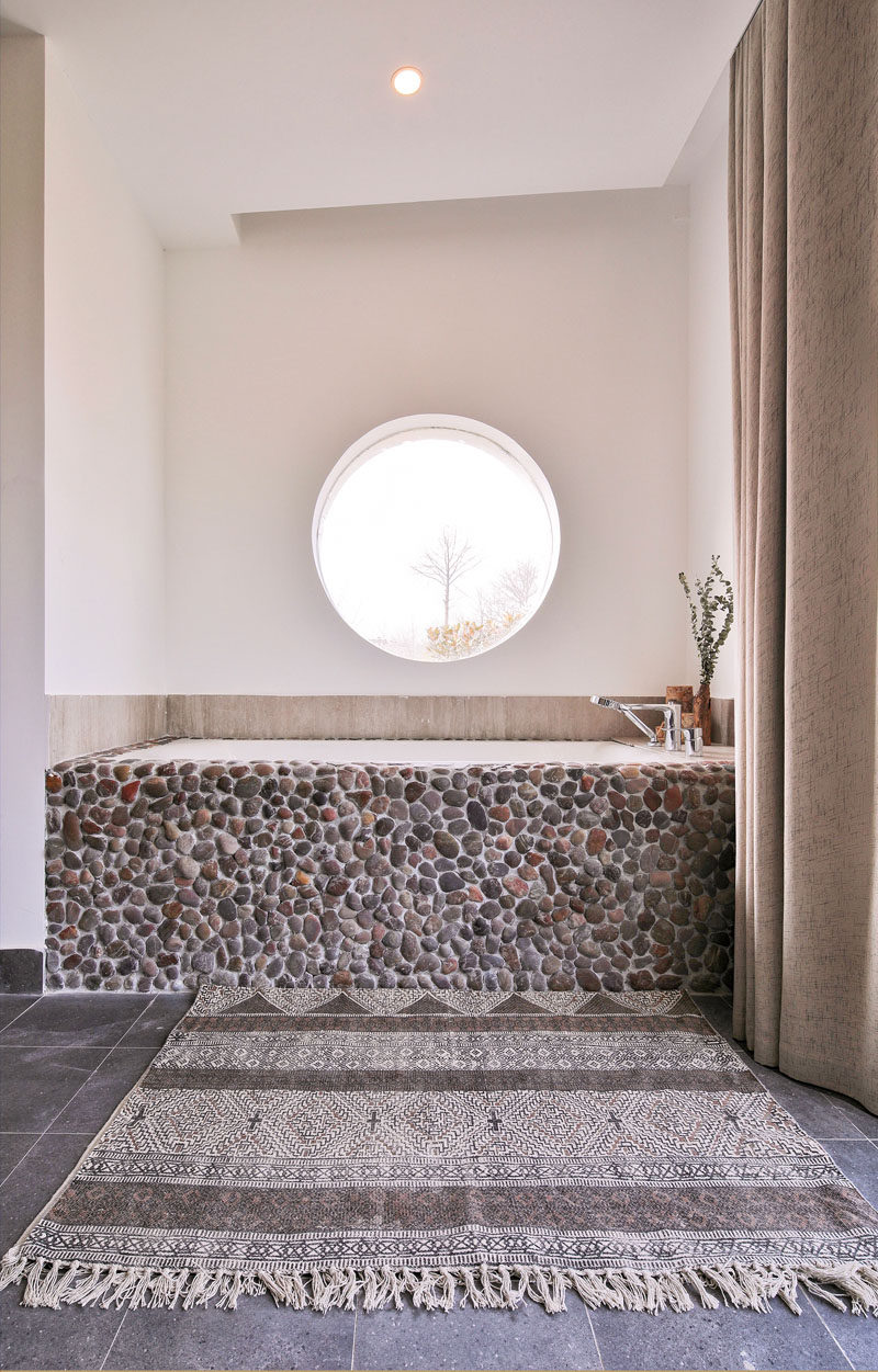 This modern bathroom features a built-in bathtub with a pebble front, that sits below a circular window. #BuiltInBathtub #BathroomDesign #Natural #Windows