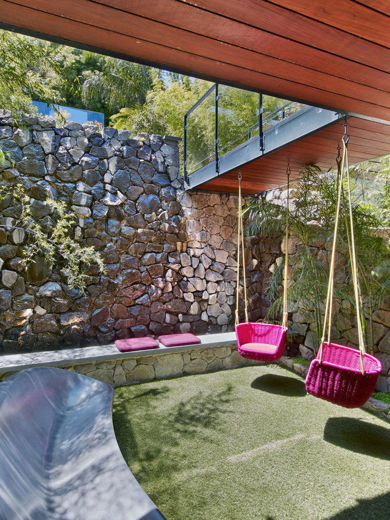 This modern house has a small grassy area with swings that hang from the bridge above, while a stone and concrete bench, and a outdoor sculptural sofa provide places to relax. #Landscaping #SmallYard #RockWall