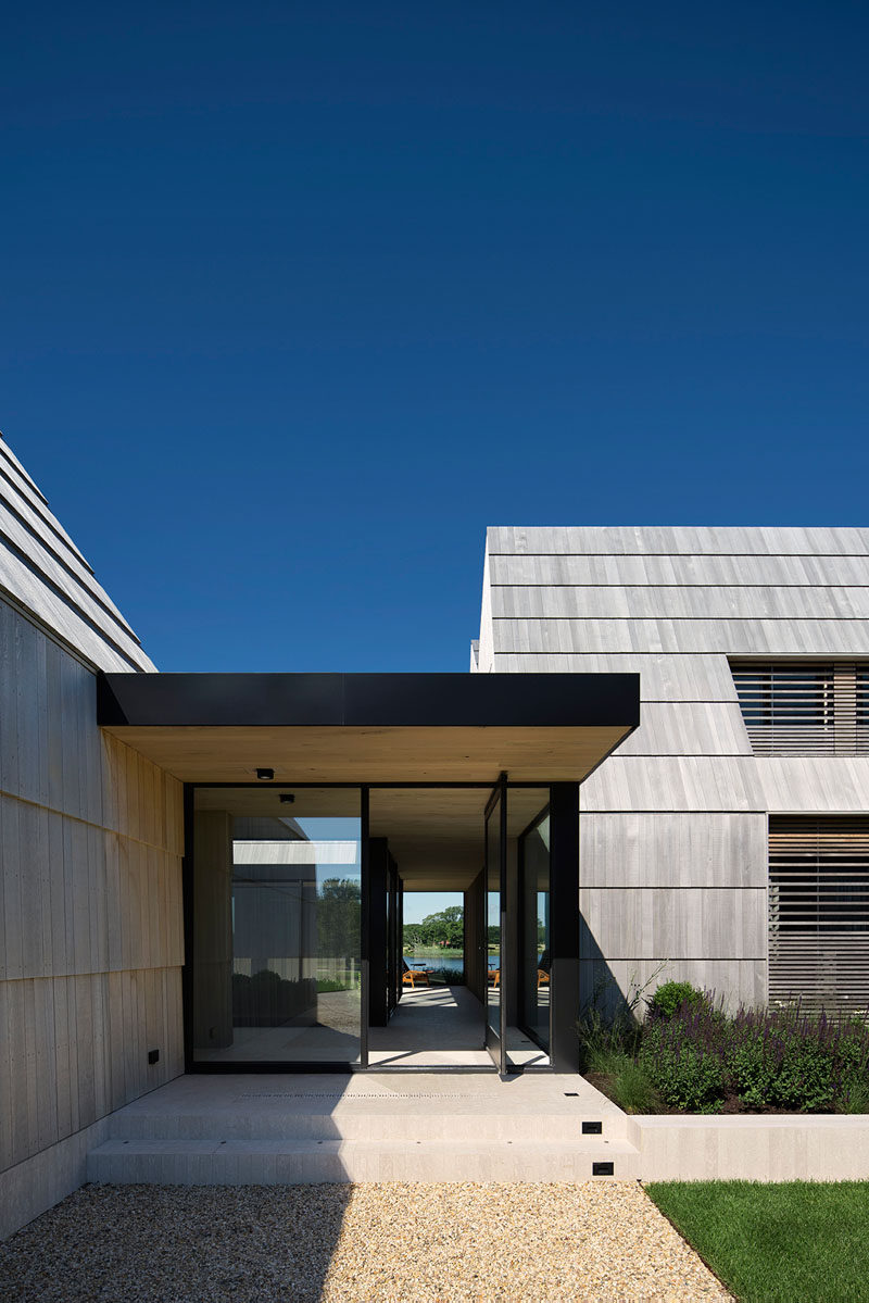 A glass pivoting front door welcomes people to this modern house that has cedar shingles. The cedar shingles have been scaled up to the size of boards to cover the roof and sidewalls, while cedar screens provide privacy and filter light. #Shingles #PivotingFrontDoor #Architecture #ModernHouse