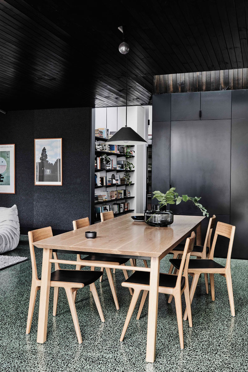 This modern house extension features a dining area furnished with a light wood table and chairs. Off to the side of the dining room is a shelving unit for the home owners book collection. #DiningRoom #Shelving #ModernDiningRoom #BlackWalls