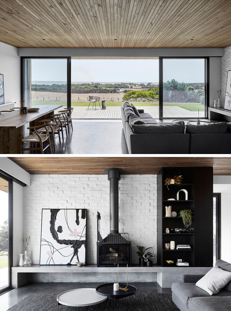 The dining and living areas of this modern house open up to a wood deck that has sweeping views of the coastline and water beyond. #ModernInteriorDesign #WhiteBrick #BlackFireplace #Deck #SlidingGlassDoors #WoodCeiling