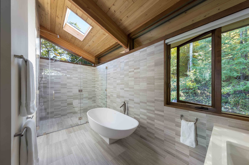 In this modern master bathroom, light colored tile and plenty of windows keep the space bright, while a glass enclosed shower is located at the end of the bathroom, with a freestanding bathtub nearby. #MasterBathroom #ModernBathroom #Windows #Skylight