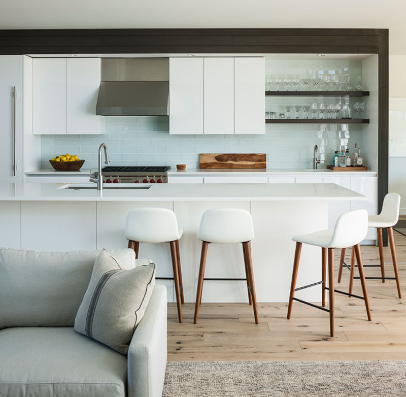 This modern kitchen has a white lacquer finish, with a glossy tropical white glass tile backsplash. The design of the kitchen island includes an overhang to allow for seating. #ModernWhiteKitchen #KitchenDesign #InteriorDesign