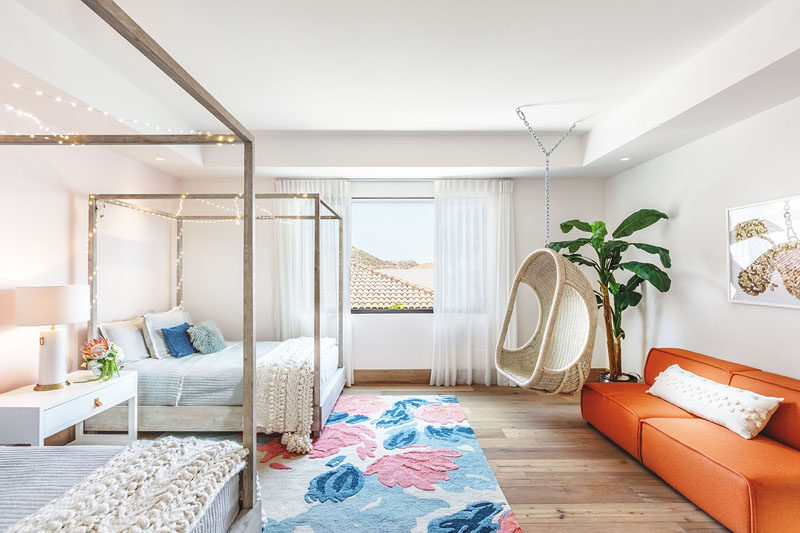 In this modern bedroom, there's two beds that have been decorated with fairy lights, and on the opposite wall there's a bright orange couch, and a woven chair hangs from the ceiling. #ModernBedroom #BedroomWithTwoBeds #InteriorDesign