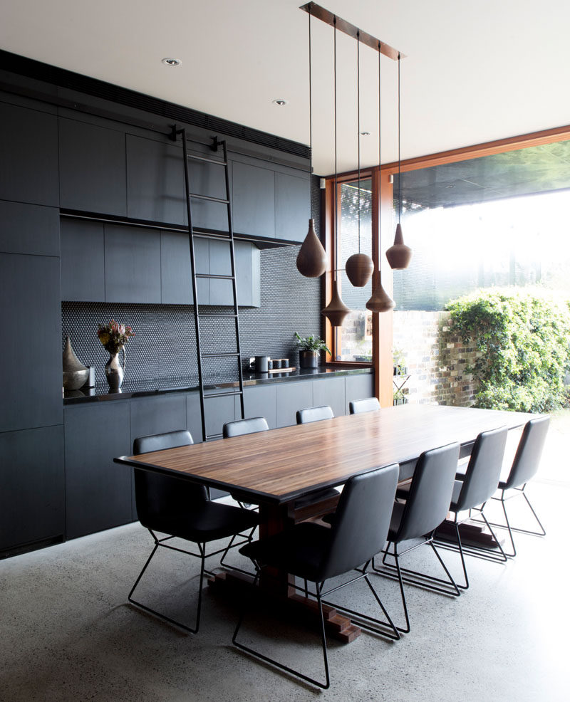 In this modern dining area, a ladder has been added to the black cabinetry to provide easy access to the upper cabinets. A collection of simple and elegant turned wood pendant lights help to anchor the dining table in the open plan interior. #DiningRoom #BlackInterior #BlackAndWood #DiningTable