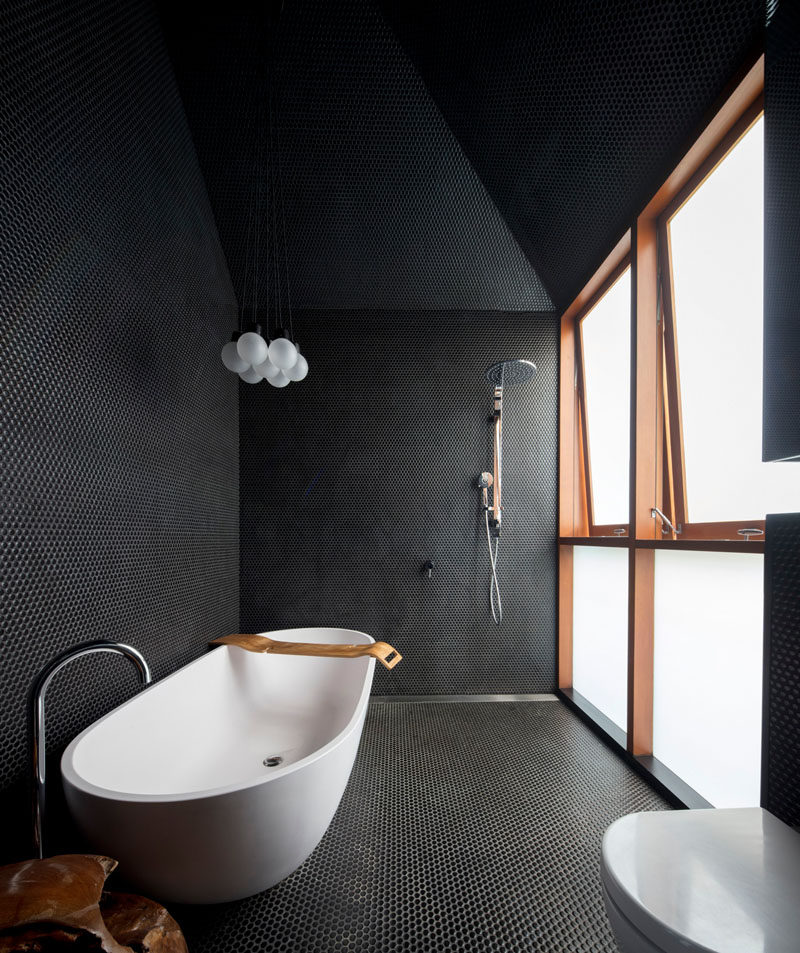Small black penny tiles cover the surfaces in this modern black bathroom, and a frosted windows provides natural light, while a white freestanding bathtub keeps things bright. #BlackBathroom #ModernBathroom #BathroomDesign