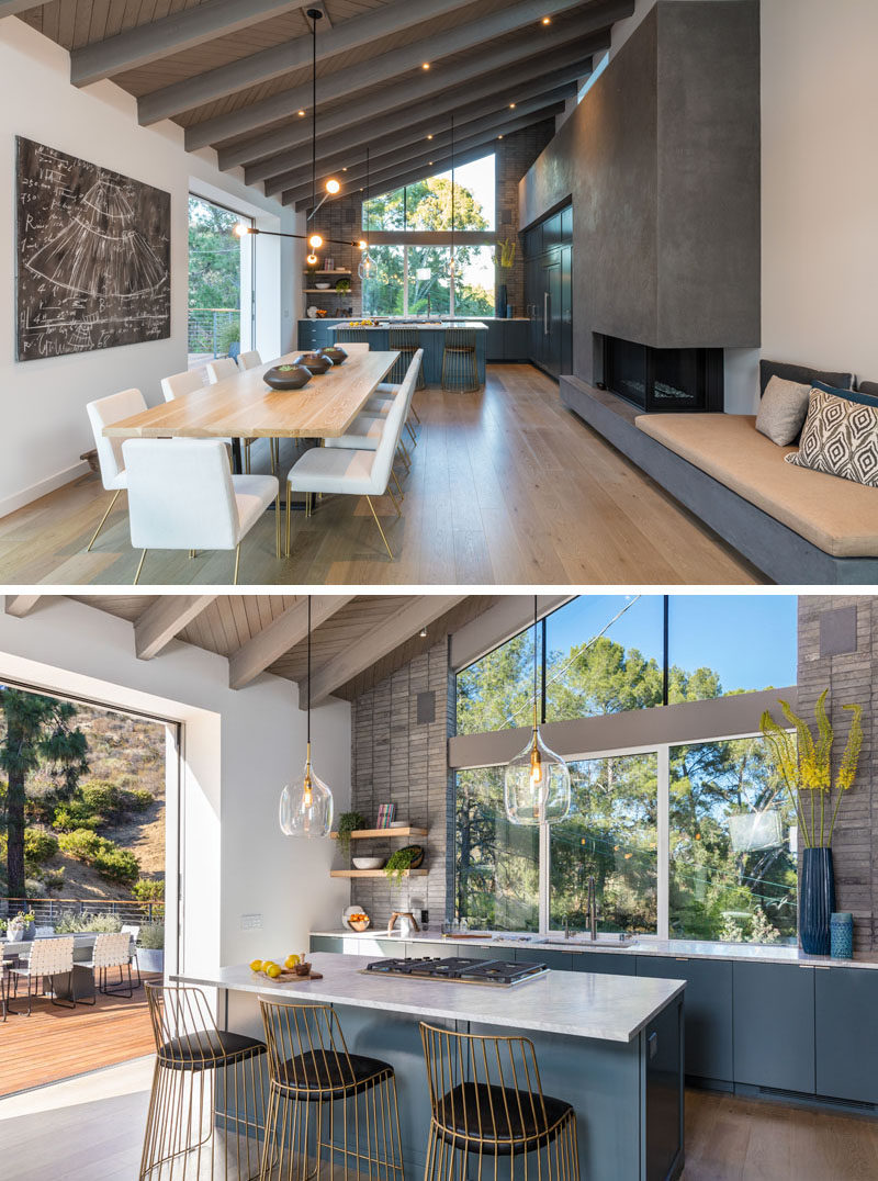 In this dining and kitchen area, a long wood dining table sits beside a built-in concrete bench and fireplace. In the kitchen deep matte blue cabinetry is topped with marble counters. #DiningRoom #KitchenDesign #BuiltInBench #BlueKitchen