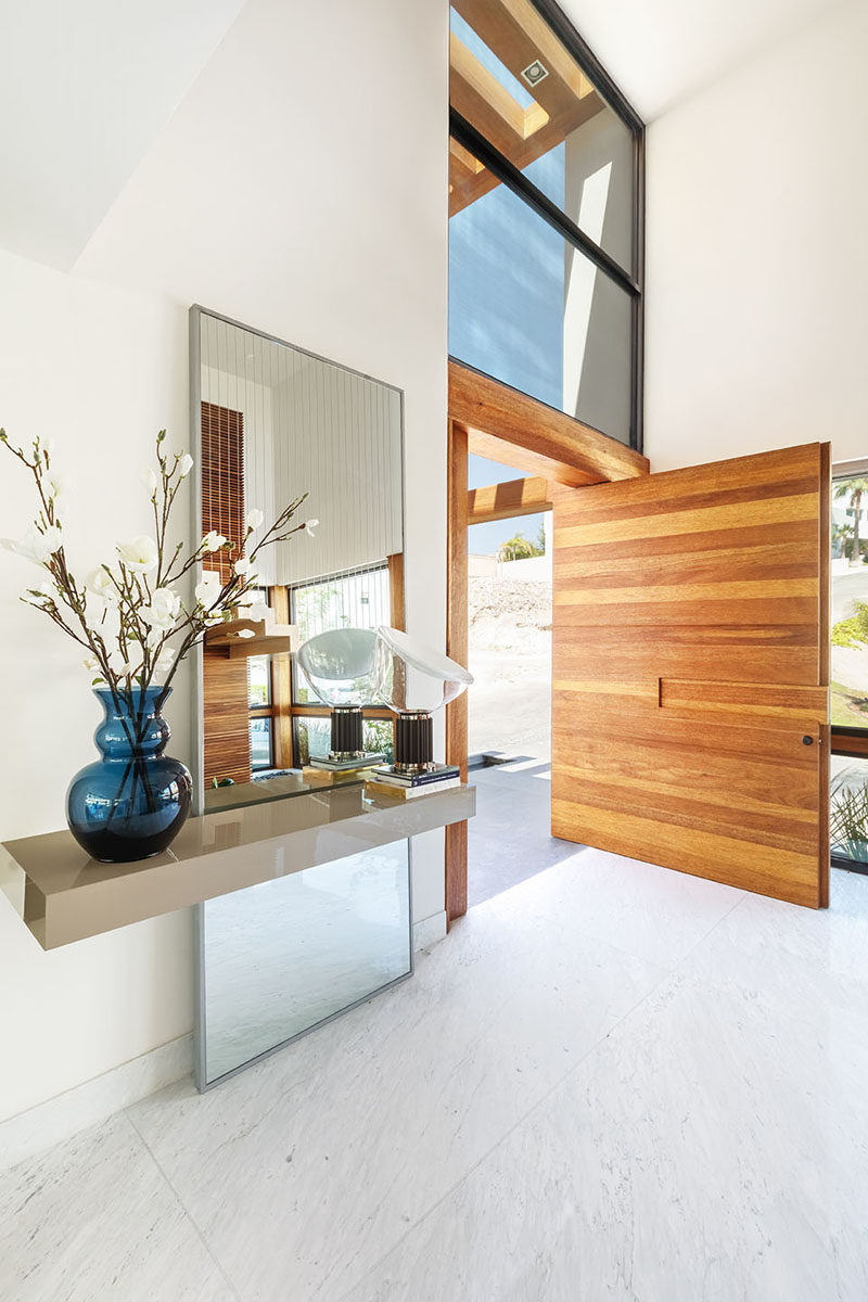 An oversized, pivoting, wood front door welcomes visitors to this modern house, and it opens up to a large double-height foyer. #WoodFrontDoor #PivotingDoor #ModernFrontDoor