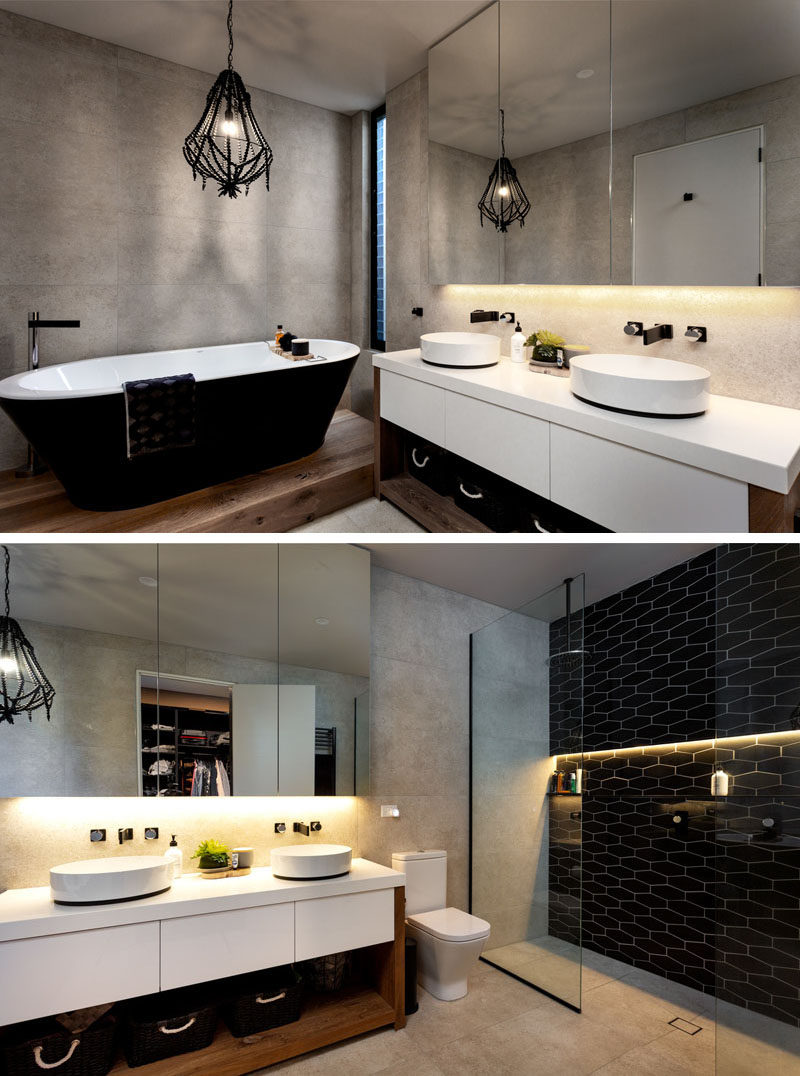 In this modern master bathroom, a black freestanding bathtub with a white interior sits below a black pendant lamp. On the other side of the vanity, a walk-in shower has a black tile accent wall with a strip of hidden lighting. #BlackBathroom #ModernBathroom #InteriorDesign