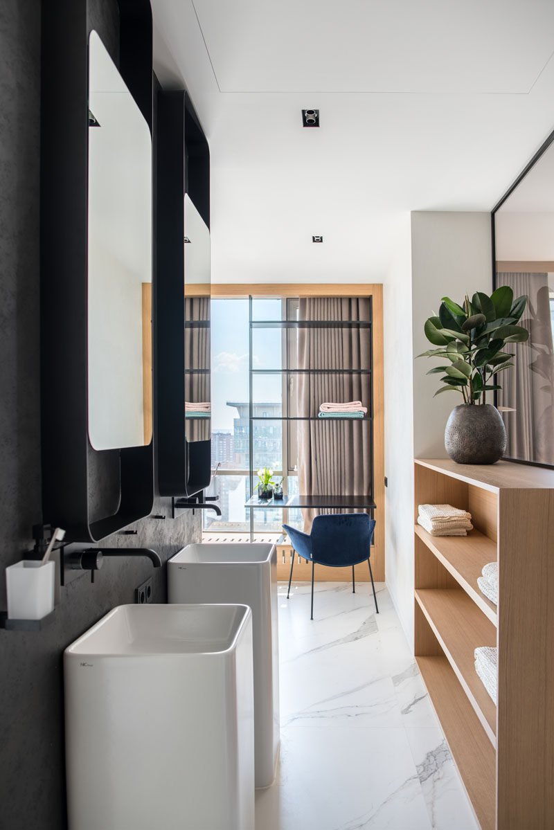 This modern ensuite bathroom features a dark accent wall, white marble tiles, wood shelving, and a desk by the window. #EnsuiteBathroom #ModernBathroom