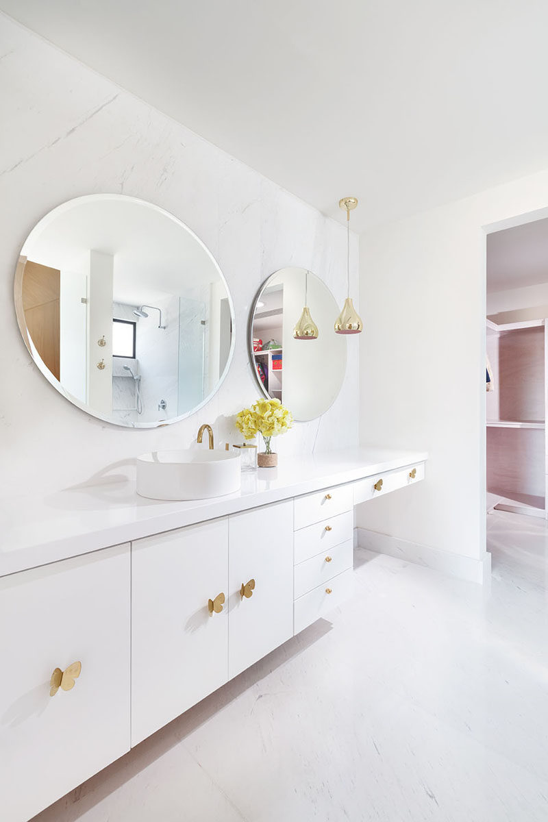 This modern white bathroom has large round mirrors, a long white vanity and a make-up area. #ModernBathroom #WhiteBathroom