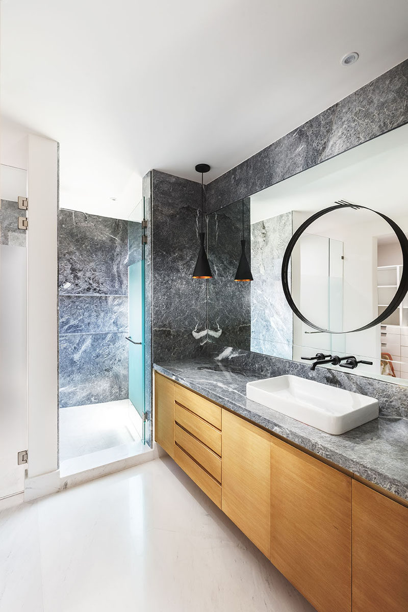 In this modern ensuite bathroom, grey stone covers the walls and is used for the counter on top of the light wood vanity. #GreyAndWoodBathroom #BathroomDesign #ModernBathroom