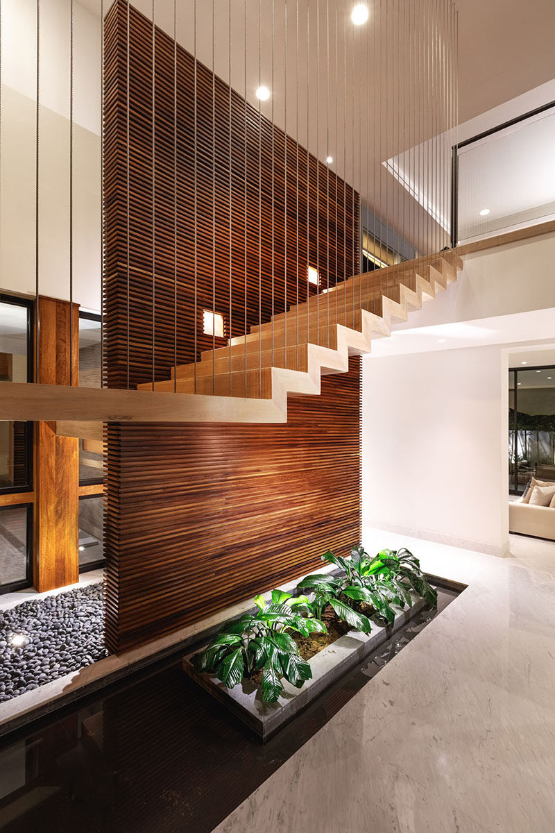 This modern staircase runs alongside a wood slat wall made from Parota. A touch of nature has been included underneath the stairs in the form of a water feature and planters. #WoodSlatWall #ModernStairs #StairDesign #InteriorWaterFeature